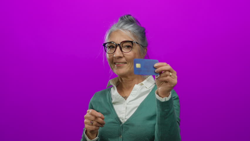 Senior woman with grey hair smiling and pointing while holding a credit card against an isolated pink background.