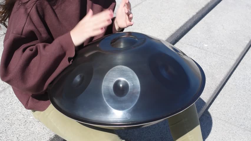 young caucasian woman playing black handpan with hands, ethnic metal drum instrument, sits on stone staircase, beautiful relaxing music for meditation, close-up view of hands