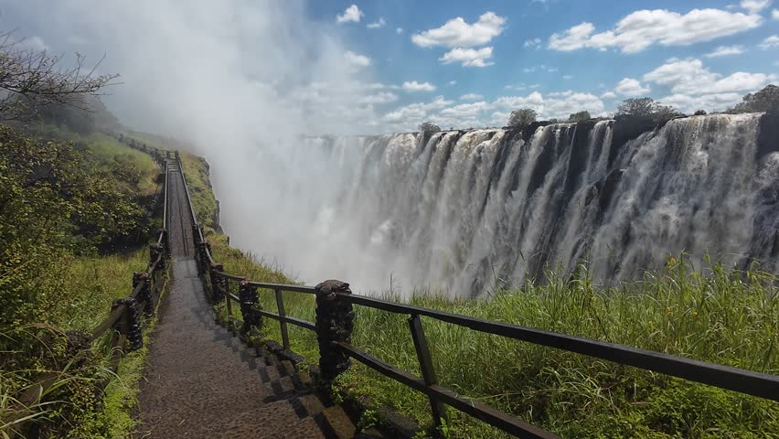 A walkway surrounded by greenery with Victoria Falls under clear sky with scattered clouds in Zimbabwe
