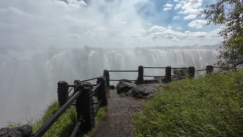 A path surrounded by greenery with Victoria Falls in the background under cloudy sky in Zimbabwe