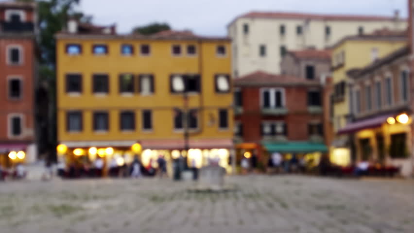 Blurred beautiful city square in Venice with souvenir shops and residential buildings, hotels illuminated in the evening in summer during tourist season at sunset.