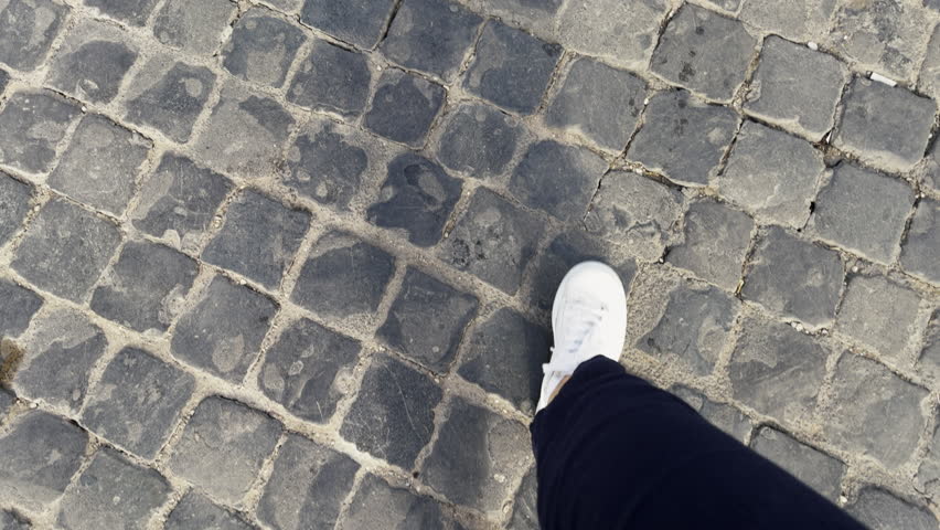 Single white sneaker on gray cobblestone pavement walk woman tourist. Person wearing dark blue pants walking. Urban exploration. City street. Shoe stepping on rough cobblestone ground