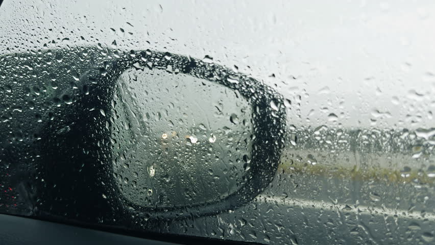 Rainy day view from car showing side mirror covered raindrops, wet road conditions creating moody travel atmosphere, driving safety theme for weather-related content
