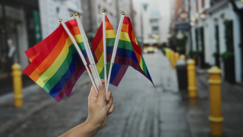 Hand holding multiple rainbow flags on a city street signifies lgbtq pride in a vibrant urban setting during a daytime event, showcasing diversity and inclusivity outdoors.