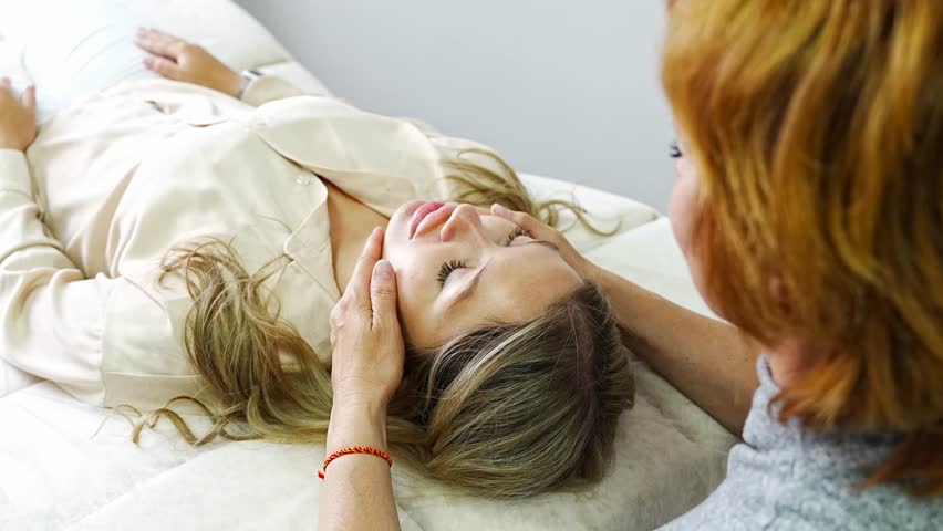 Female practitioner performs facial lifting technique on young woman lying on couch. Concept of energy healing, face therapy and alternative wellness practice from Access Bars. High quality 4k footage