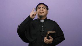 Smiling asian priest holding a bible pointing up with a joyful expression against a purple background - Powered by Shutterstock - Get 15% off with code: PIKWIZARD15