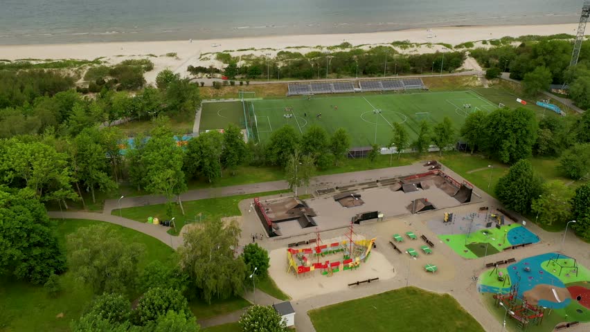 Aerial view of a recreational area featuring a playground, sports fields, and beach in the background. Lush greenery surrounds the facilities, creating a vibrant outdoor space.