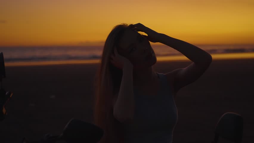 Cool girl posing with motorcycle at sunset, woman with bike on the beach in dark setting. Feeling freedom and independence, having moto trips and exciting adventures.