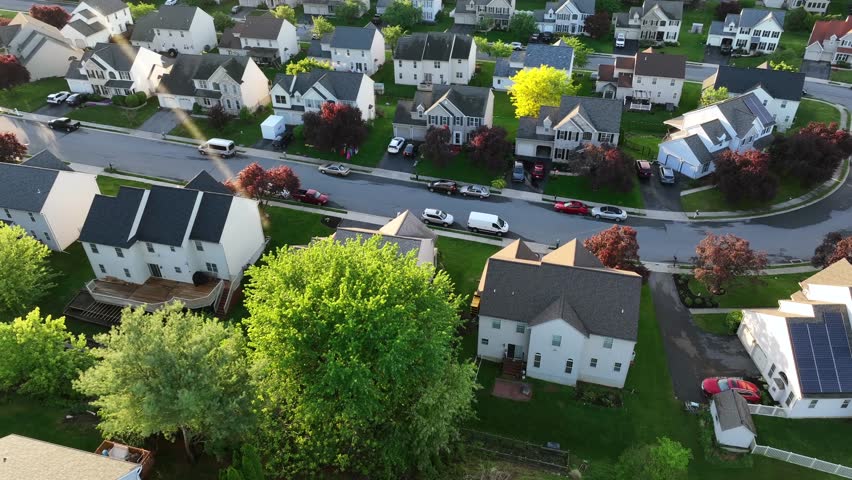 Aerial approaching shot of single Family houses with colored trees in spring. Calm evening sunset in suburb. Top down. Row of homes with parking cars on street. Quiet neighborhood of United States.