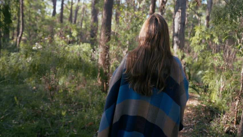 Young Caucasian woman walks thoughtfully along a path in a peaceful forest. Sunlight filters through the trees, creating a serene atmosphere for contemplation.