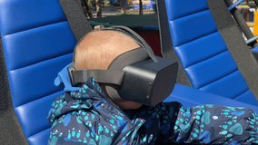 A young child, wearing a virtual reality headset and a patterned blue jacket, sits intently in a blue padded seat with a black hand control, appearing to be on a ride or in a gaming setup - Powered by Shutterstock - Get 15% off with code: PIKWIZARD15