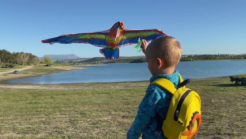 A young child, seen from behind and wearing a bright yellow dinosaur-themed backpack, stands by the calm waters of a lake, holding a large, colorful kite, ready to launch it under a clear blue sky.