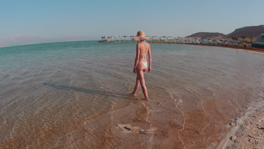 Slender girl in a bathing suit walks along the sandy sea beach of the Dead Sea in Israel. Girl on vacation. Conceptual travel and vacations.