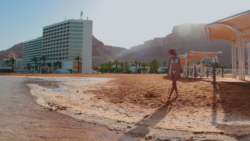Slender girl in a bathing suit walks along the sandy sea beach of the Dead Sea in Israel. Girl on vacation. Conceptual travel and vacations.