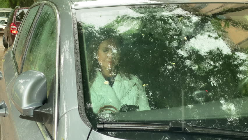 A woman is seen through the rain-splattered windshield of a car, appearing to be in the driver