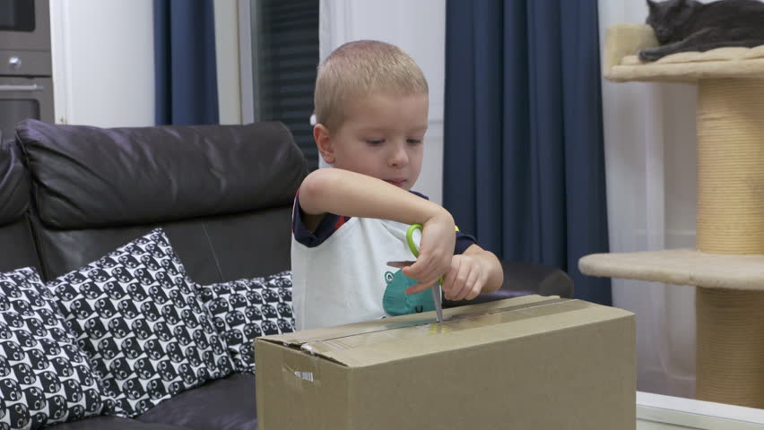 A young boy with blond hair carefully opens a cardboard box using a pair of green-handled scissors. He's indoors at home, standing in front of a black leather couch adorned with patterned pillows.