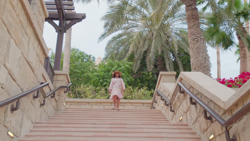 young woman climbing down the stairs in slow motion, with old buildings, European architecture, trees in background, she is wearing a floral dress and hat, pro-res video	