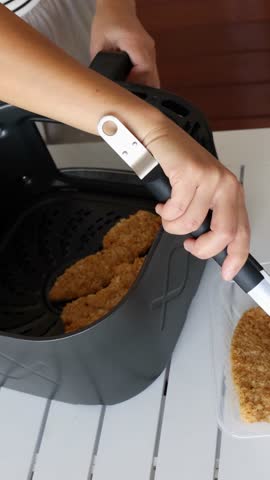 A person uses tongs to place breaded chicken tenders into an air fryer on a white table