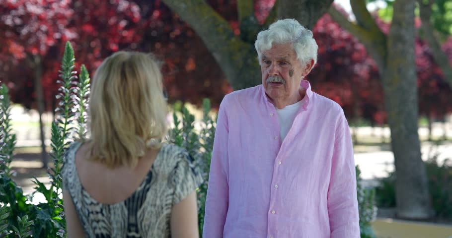 Elderly man in a pink shirt emotionally explaining something to a woman in a patterned dress while standing in a green park with blooming flowers