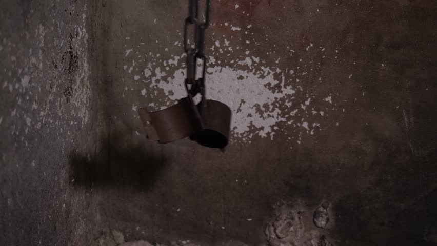 Close-up of an old rusty chain with handcuffs hanging on the gray dirty wall of the prison. Various scary tools in the torture room. The room of horrors. An old prison room.