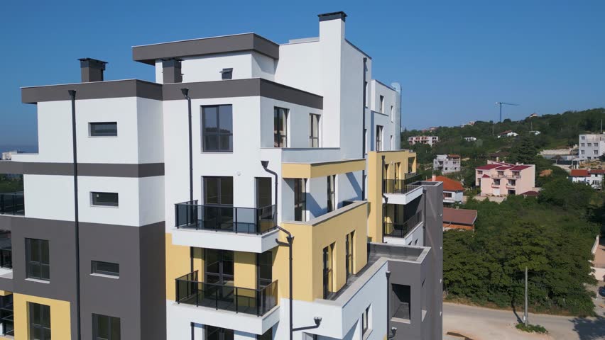 Aerial drone shot orbiting a modern residential building with clean architecture, large windows, and balconies, set in an urban environment under clear blue sky