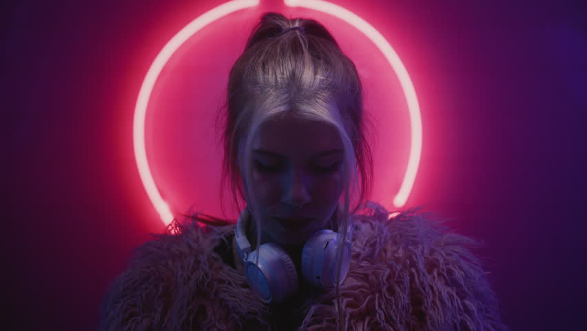 Young woman DJ looking at the camera in the neon light of the night club. Portrait of a beautiful artist girl close-up. Lady in multicolor glow. Face of a female, put on white headphones.