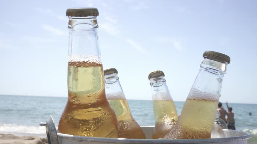Chilled beer bottles nestled in frosty ice bucket, surrounded by golden sand, sparkling ocean waters stretching across sunny tropical horizon