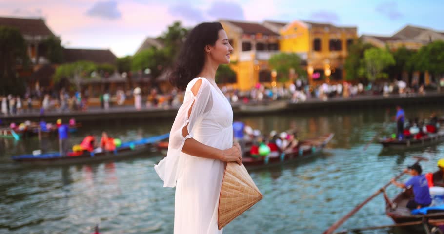 A serene slow-motion video showing a woman in a flowing white dress looking at the river in Hoi An, Vietnam, from behind. Ideal for use in travel, fashion, cultural or lifestyle media projects.