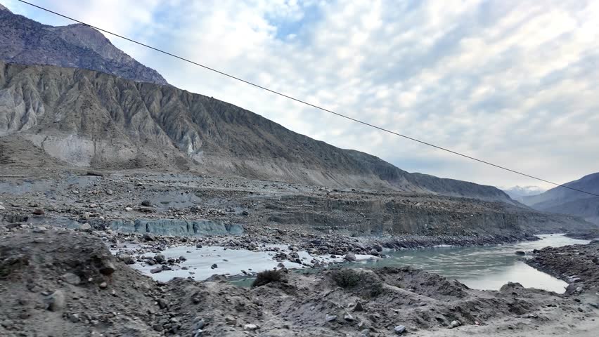 A tranquil river winds through a rugged mountain valley, framed by majestic cliffs under a cloudy sky. This serene landscape invites exploration and adventure.