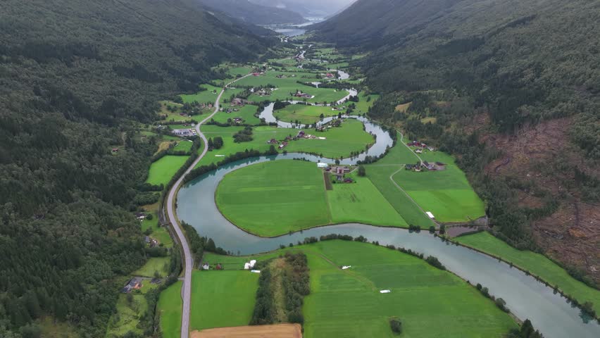 A drone flying on Stryn River in green land with scattered houses, surrounded by mountains under cloudy sky in Stryn, Norway