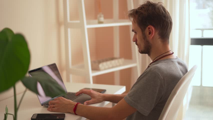 A young man sits at a clean desk in a bright room, intently typing on his laptop. The space blends productivity and modern workspace aesthetics, highlighting contemporary work environments