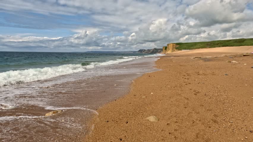 Slow Motion footage of the waves crashing up onto a beach on the Jurassic Coast, Dorset. The footage is low and moves backwards with amazing cliffs in the background and a sky full of clouds above.