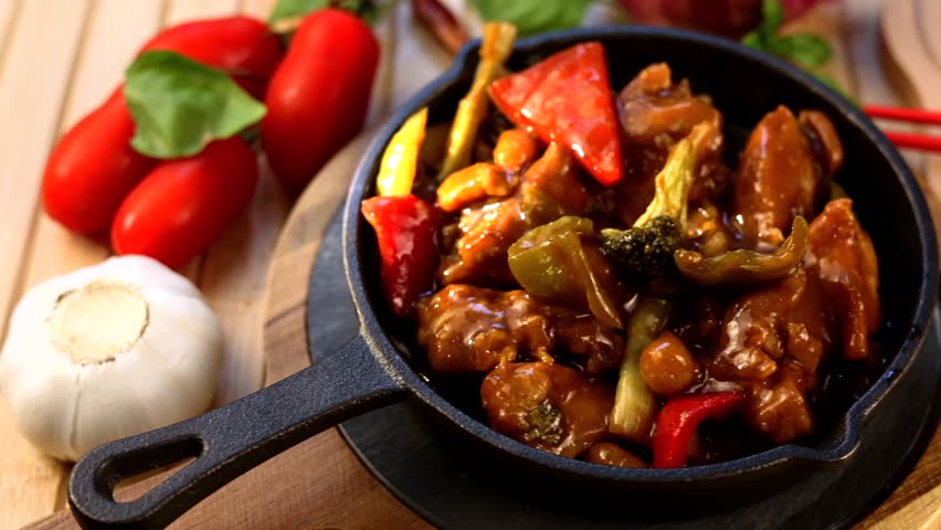 Cast iron skillet holding sizzling chicken cashew stir fry, garnished with cherry tomatoes, garlic, basil leaves, red chopsticks resting on rustic wooden surface