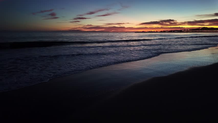Gimbal wide panning shot of the painted sky with reflecting light in the gentle waves during magic hour on East Beach in Santa Barbara, California. 4K