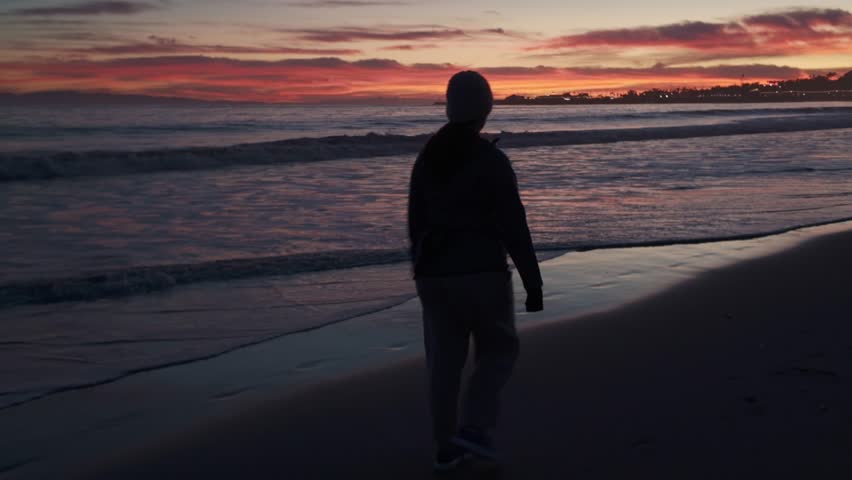Gimbal close-up shot following a silhouetted woman walking on a beach during magic hour in Santa Barbara, California. 4K