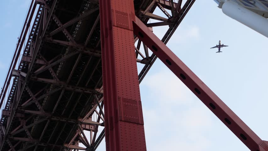 Low-angle view of the 25 de Abril Bridge in Lisbon, Portugal, with a passenger airplane flying overhead. Red steel structure and visible rivets add to the industrial composition and urban mood.
