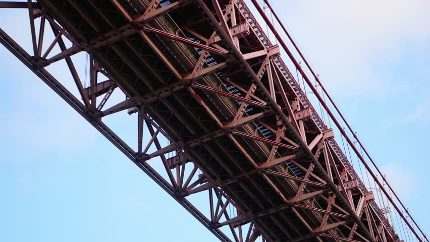 Low-angle close-up views of the 25 de Abril Bridge in Lisbon, Portugal, highlight the industrial urban mood aesthetic and bold geometric composition of the red steel structure. 