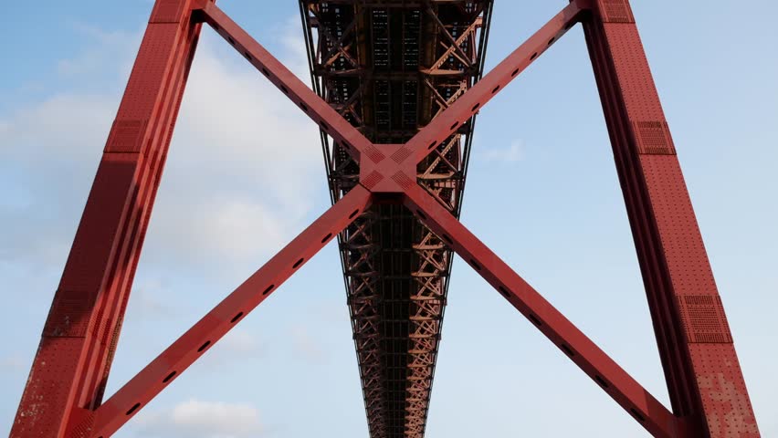 Low-angle close-up views of the 25 de Abril Bridge in Lisbon, Portugal, highlight the industrial urban mood aesthetic and bold geometric composition of the red steel structure. 