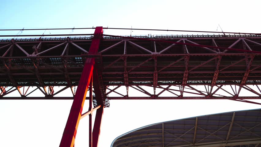 Low-angle close-up views of the 25 de Abril Bridge in Lisbon, Portugal, highlight the industrial urban mood aesthetic and bold geometric composition of the red steel structure. 