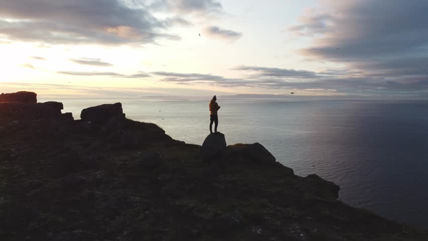 Edge of the world, on the final cliff plunging into the ocean along Icelands Snaefellsnes Peninsula. Man stands in awe, gazing into the depths of an ocean as the sky burns with a breathtaking sunset.