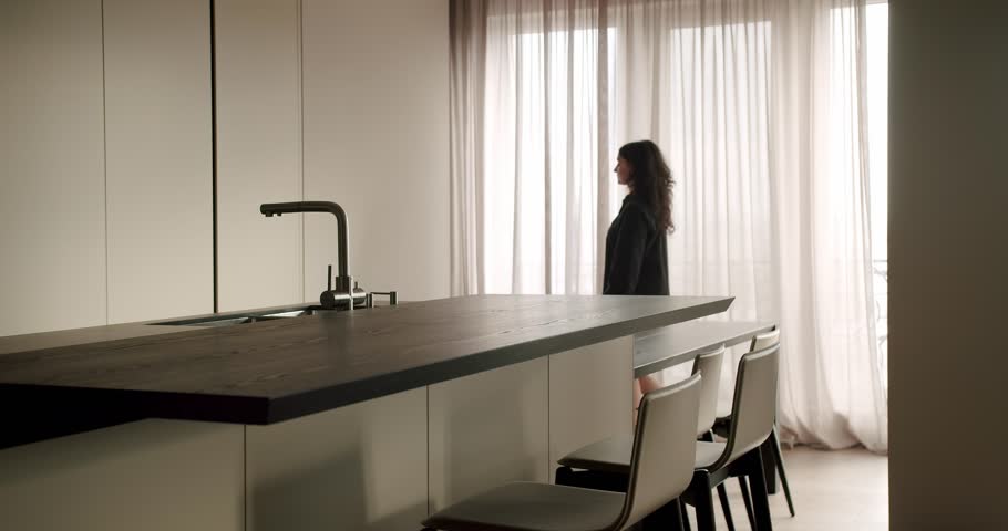 A thoughtful woman in a dark blazer peers out a bright window, standing in a sleek, modern kitchen with a stylish design. Elegant Woman in Modern Kitchen Gazes Out The Window