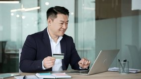 Asian businessman enters a credit card data on laptop sitting at workplace in business office. Entrepreneur in formal suit shopping online, happy about a successful transaction, e-commerce, e-shopping - Powered by Shutterstock - Get 15% off with code: PIKWIZARD15