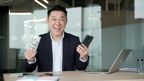 Happy asian businessman holding fan of cash money dollar bills and mobile phone sitting in a business office looking at camera. Male is satisfied with earning money using application on his smartphone - Powered by Shutterstock - Get 15% off with code: PIKWIZARD15