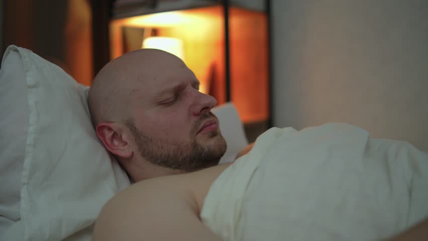 Man wakes up in the middle of the night, writhes in pain and touches his neck. Concept of neck pain due to incorrect sleeping position and bad pillow