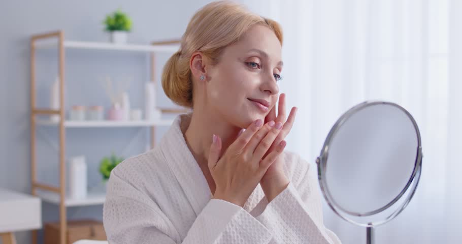Middle aged blonde woman looking at her reflection in mirror, touching soft facial skin, satisfied with its condition after beauty treatments, wearing white bathrobe, posing indoor. 