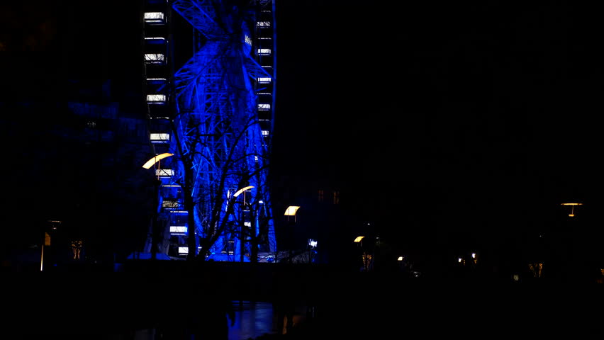Ferris wheel rotating at night with blue lights. Massive ferris wheel revolving against dark nighttime sky, glowing with vivid blue illumination and creating mesmerizing visual spectacle