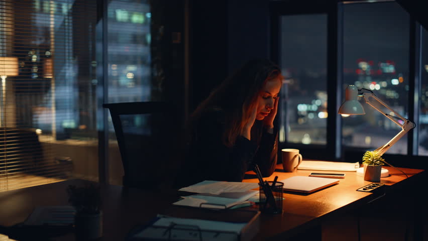 Fatigued employee working overtime in dark modern office interior closeup. Tired manager staying alone during late shift. Exhausted professional resting solo at nighttime desk at corporate workspace