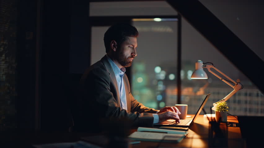 Late worker typing computer taking coffee sip sitting office desk closeup. Bearded confident corporate employee looking screen flipping documents at workplace. Serious businessman doing laptop work 