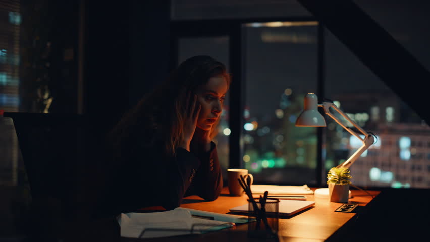 Weary office worker touching forehead remaining long hours inside corporate workspace closeup. Drained executive resting solo at nighttime desk. Burned out professional finishing task holding head