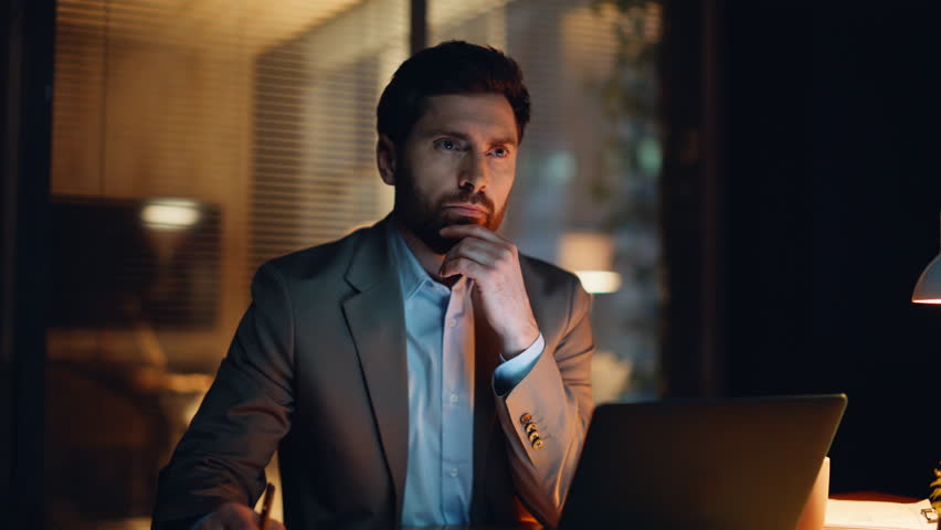 Serious businessman browsing computer writing notes at cozy dark flat closeup. Focused bearded man looking laptop screen texting email at home. Overworked handsome ceo typing keyboard at night house 